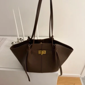 Brun axelväska med gulddetaljer - Snygg brun axelväska i skinn med elegant gulddetalj framtill. Väskan har en stilren design med långa axelremmar och en praktisk stängning. Perfekt för att bära dina dagliga nödvändigheter med stil.