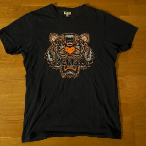 Marinblå t-shirt med tigertryck från Kenzo - Snygg marinblå t-shirt från Kenzo med ett coolt tigertryck i orange och ljusblått på framsidan. Perfekt för att ge din outfit en edgy touch. Kortärmad och tillverkad i mjukt material för extra komfort. Säljer för den blivit för liten tyvärr..