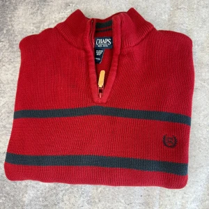 Röd tröja med dragkedja från Chaps - Säljer en snygg röd tröja från Chaps med dragkedja och svarta ränder. Tröjan är tillverkad i 100% bomull och har långa ärmar. Perfekt för en stilren look.