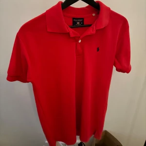 Röd pikétröja från Ralph Lauren - Säljer en klassisk röd pikétröja från Ralph Lauren. Tröjan är tillverkad i 100% bomull och har en broderad logotyp på bröstet. Perfekt för en stilren och avslappnad look.