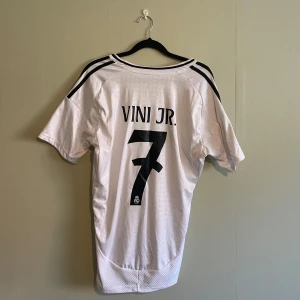 Real Madrid fotbollströja från Adidas - Säljer en vit fotbollströja från Adidas med korta ärmar. Tröjan har svarta detaljer och klubbmärke på bröstet. Den har även texten 'VINI JR.' och nummer 7 på ryggen. Perfekt för fotbollsfans!