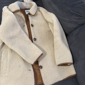 Beige teddyjacka från Zara - Säljer en mysig beige teddyjacka från Zara med bruna detaljer. Jackan har knappar framtill och en klassisk krage. Perfekt för kyliga dagar när du vill hålla dig varm och stilren. Storlek S men passar även för M