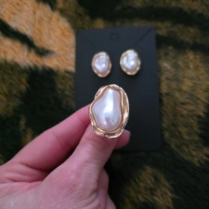 Guldpläterade örhängen och ring med pärlor - Snyggt set med guldpläterade örhängen och matchande ring, båda med stora vita pärlor. Perfekt för att ge en elegant touch till din outfit. Örhängena är i en klassisk design och ringen har en justerbar storlek.