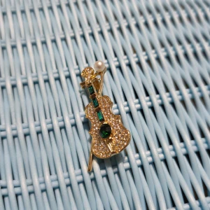 Glittrig violinbrosch med pärldetalj - Elegant brosch i form av en violin, dekorerad med glittrande stenar och en vit pärla. Den har gröna detaljer som ger en lyxig känsla. Perfekt accessoar för att ge en unik touch till din outfit.