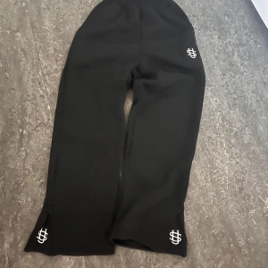 Svarta byxor med dragkedja och broderad logga - Snygga svarta byxor med broderad logga på benen. Byxorna har dragkedjor vid bensluten för en cool detalj. Perfekta för en stilren look.