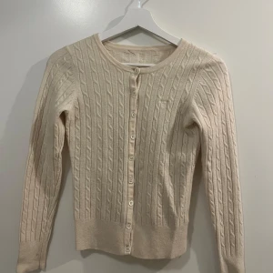 Beige kabelstickad kofta från kant - Säljer en stilren beige kabelstickad kofta med knappar framtill. Den har en rund halsringning och långa ärmar, perfekt för kyligare dagar. Koftan är både mjuk och bekväm, vilket gör den till ett mångsidigt tillskott i garderoben.