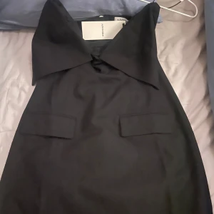 Svart klänning från Adoore - Elegant svart klänning från Adoore med en stilren design. strapless och dekorativa fickdetaljer framtill. Den är axelbandslös och har en dragkedja baktill för enkel påklädning. Orginalpris 1600kr, helt ny. Passar s/m