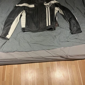 Svart och vit mc skinnjacka från Held - Säljer en cool svart och vit skinnjacka från Held. Jackan har långa ärmar och dragkedja framtill. Den är designad med vita paneler och svarta detaljer, vilket ger en snygg kontrast. Perfekt för motorcykelentusiaster!