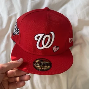 Röd keps från New Era - Snygg röd keps från New Era med broderad 'W' och hjärta på framsidan. Sidan har texten 'Love & Hustle = Game' och World Series-märke. Perfekt för sportfantaster!