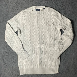 Gant tröja  - Säljer en stilren grå kabelstickad tröja från Gant. Tröjan har långa ärmar och en rund halsringning. Perfekt för kyligare dagar med sin klassiska design och mjuka material.