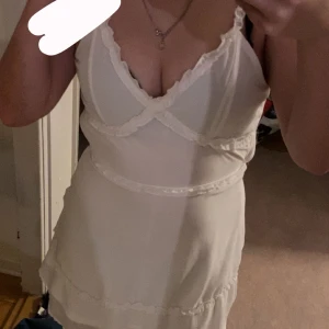 Vit klänning med spetsdetaljer - Säljer en söt vit klänning med spetsdetaljer och smala axelband. Klänningen har en omlott-design och är perfekt för varma dagar.