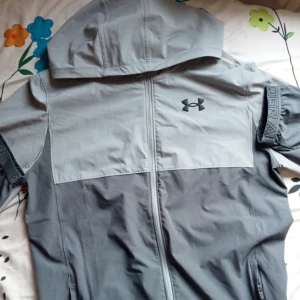 Grå vindjacka från Under Armour - Säljer en grå vindjacka från Under Armour med huva och dragkedja. Jackan är XL i barn storlek och M i vuxen storlek.
