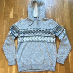 Grå stickad hoodie med med päls luva  - Säljer en mysig grå stickad hoodie med ett snyggt mönster i svart och vitt över bröstet och ärmarna. Tröjan har långa ärmar och en praktisk huva med päls. Perfekt för kyliga dagar!
