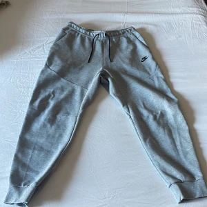 Grå mjukisbyxor från Nike - Säljer ett par grå mjukisbyxor från Nike med elastisk midja och snörning. Byxorna har sidofickor och en liten Nike-logga på ena benet. Perfekta för en avslappnad stil. Även nästan helt oanvända och nypris 1200 kr