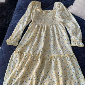 Blommig klänning, strl 150 - Säljer en söt blommig klänning från Shein i ljusgult och grönt. Klänningen har långa ärmar och en smockad överdel som ger en fin passform. Perfekt för vår och sommar med sitt blommiga mönster och volangdetaljer.