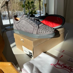 Grå sneakers från Christian Louboutin - Helt nya snygga grå sneakers från Christian Louboutin med ikoniska röda sulor och nitar på tån. Skorna har snörning och är tillverkade i mocka, vilket ger en lyxig känsla. Perfekta för den som vill sticka ut med stil.