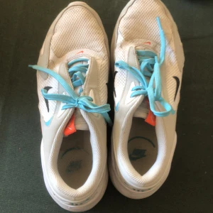 Vita Nike Air Max med blå snören - Säljer ett par vita Nike Air Max med ljusblå snören och orange detaljer. Skorna har en synlig luftkudde i sulan och en svart Nike-logga på sidan. Perfekta för en sportig look!