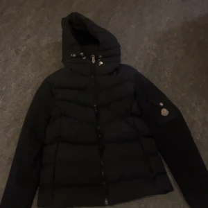 Svart dunjacka med huva - Säljer en stilren svart dunjacka från Moncler med huva och dragkedja. Jackan har långa ärmar och en quiltad design. Perfekt för våren.