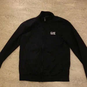 Svart jacka från Emporio Armani - Snygg svart jacka från Emporio Armani med dragkedja och EA7-logga på bröstet. Jackan har en stilren design med långa ärmar och en hög krage. Perfekt för en cool och avslappnad stil.