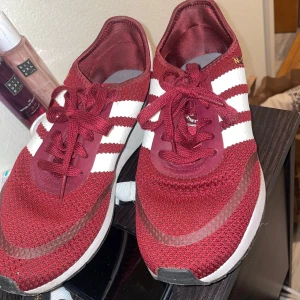 Vinröda adidas sneakers  - Snygga vinröda adidas sneakers med vita ränder och snörning. Perfekta för en avslappnad stil. Skorna har en bekväm sula och är tillverkade i syntetmaterial.