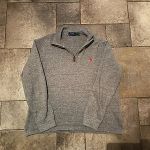 Ralph Lauren Quarter Zip Medium - Säljer en stilren grå Quarter/Zip från Polo Ralph Lauren med en halv dragkedja och det klassiska röda logotypbroderiet på bröstet. Perfekt för en avslappnad men snygg look. Storlek Medium och i väldigt gott skick.