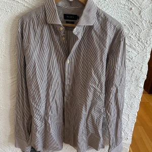 Randig skjorta från Riley - Snygg randig skjorta från Riley i slim fit. Skjortan är brun och vit med långa ärmar och knappar framtill. Tillverkad i 100% bomull för en bekväm känsla. Perfekt för en stilren look.