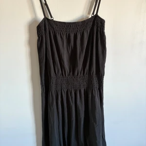 Svart axelbandslös klänning - Snygg svart klänning med smockdetaljer och smala axelband. Perfekt för en stilren look. Klänningen har en luftig passform och är gjord i ett lätt material. Väldigt gammal men knappt använd. Strl 34