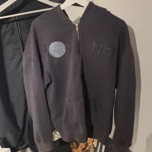 Svart hoodie från Broken Planet - Säljer en svart hoodie från Broken Planet med dragkedja och coolt planetmotiv på bröstet. Baksidan har ett unikt mönster som ger en extra touch. Perfekt för dig som vill ha en stilren men ändå iögonfallande look.