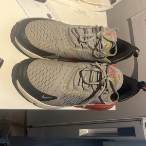 Nike Air Max 270 i grå och svart - Säljer ett par Nike Air Max 270 sneakers i grått och svart med orange detaljer. Skorna har en stor synlig Air-enhet i hälen och snörning framtill. Perfekta för en sportig look.