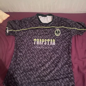 Svart och grå fotbollströja från Trapstar - Säljer en cool fotbollströja från Trapstar i svart och grått med ett unikt mönster. Tröjan har korta ärmar och en gul detalj på bröstet samt siffran 22 på ryggen. Perfekt för dig som vill sticka ut på planen!