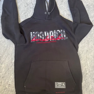 Svart hoodie från Hoodrich - Säljer en svart hoodie från Hoodrich med tryck i vitt och rött på både fram- och baksidan. Hoodien har en stor ficka framtill och justerbar huva med snören. Perfekt för en casual look.