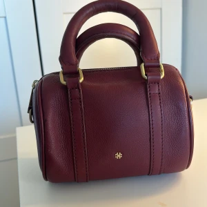 Vinröd handväska från DAY ET - Snygg vinröd handväska från DAY  ET i skinn med guldfärgade detaljer. säljs på grund av att den sällan används💗