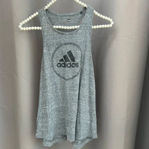 Grå ärmlös linnetopp från Adidas - Säljer en grå ärmlös linnetopp från Adidas med deras ikoniska logga framtill. Perfekt för träning eller en avslappnad dag. Tillverkad i mjukt och bekvämt material🩶