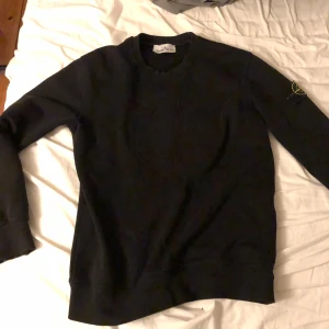Svart tröja från Stone Island - Säljer en stilren svart tröja från Stone Island med deras ikoniska logga på ärmen. Tröjan har långa ärmar och en rund halsringning. Perfekt för en casual look. Skador längst ut vid händerna.