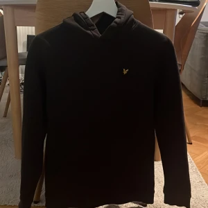 Svart hoodie junior från Lyle & Scott - Säljer en stilren svart hoodie från Lyle & Scott Junior. Tröjan har en klassisk design med en liten logga på bröstet. På fickan på magen finns en liten fläck som knappt syns. Se bild. 
