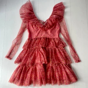 Klänning med volanger - Säljer en romantisk rosa klänning med blommigt mönster och volanger. Klänningen har mesh långa ärmar och her snörning baktill + dragkedja. Perfekt för en speciell kväll elller kommande studentfester!