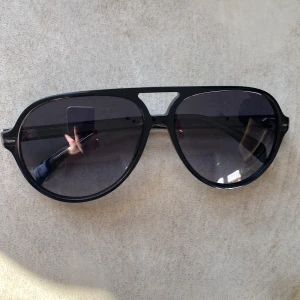 Svarta solglasögon från David Beckham - Snygga svarta solglasögon från David Beckham med en klassisk aviator-stil. De har en stilren design med grå-tonade linser. Nypris ca 2000kr, lapp/streckkod finns kvar på baksidan av fodralet. Hör av er vid minsta fundering.