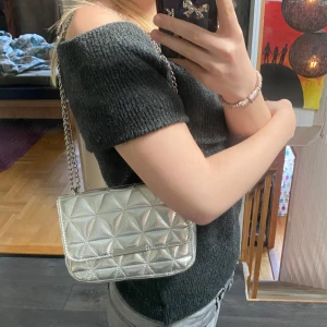 Silverfärgad quiltad axelväska - Snygg silverfärgad axelväska med quiltat mönster och kedjerem.  Kedjan går att ha som på bilden eller cross body lång. Väskan har ett fack på insidan för småsaker. 🤍