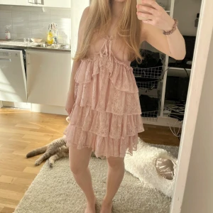 Rosa spetsklänning med volanger - Charmig rosa klänning i spets med flera lager av volanger. Klänningen har axelband och en söt knytning framtill. Perfekt för en romantisk look.