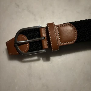 Svart flätat bälte med bruna detaljer - Snyggt svart flätat bälte med bruna läderdetaljer och silverfärgat spänne. Perfekt för att ge en stilren touch till din outfit. Bältet har en klassisk design med en modern twist.