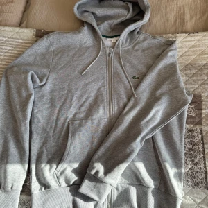 Grå hoodie från Lacoste - Riktigt fräsch Lacoste zip i storlek L. Använd under en kväll så i princip helt ny! Nypris ca 1500kr. 