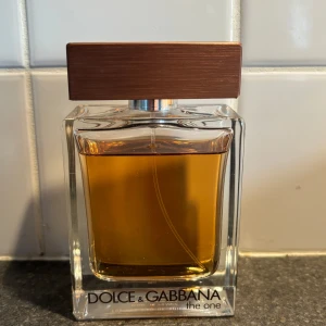 Dolce & Gabbana The One - Säljer en elegant herrparfym från Dolce & Gabbana, The One. Flaskan har en stilren design med en fyrkantig form och en brun kork. Doften är varm och kryddig, perfekt för den som vill ha en sofistikerad och tidlös parfym.