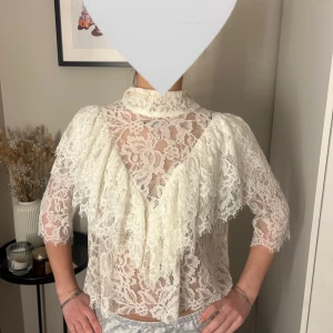 Vit spetsblus - Unik Elegant vit spetsblus med hög krage och trekvartsärmar. Blusen har ett vackert volangmönster framtill och är perfekt för fest. Fåtal använd. 