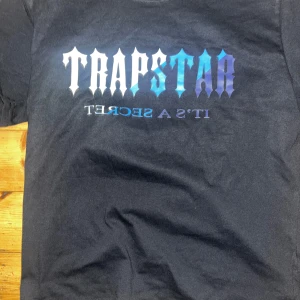 Svart t-shirt från Trapstar - Säljer en svart t-shirt från Trapstar med ett coolt tryck i blått och vitt på framsidan. T-shirten har korta ärmar och en klassisk passform. Perfekt för en avslappnad stil.