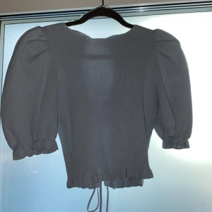 Grå ribbad blus med puffärmar - Säljer en stilren grå ribbad blus med puffiga trekvartsärmar och volangdetaljer vid ärmslut och nederkant. Blusen har en knytning i midjan för en justerbar passform. Perfekt för en chic och bekväm look.