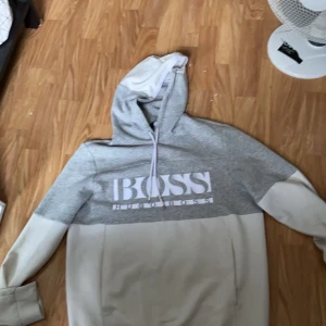 Grå och vit hoodie från Hugo Boss - Snygg grå och vit hoodie från Hugo Boss med stor logga på bröstet. Tröjan har en justerbar huva och långa ärmar. Perfekt för en avslappnad stil. Ny pris 2000kr