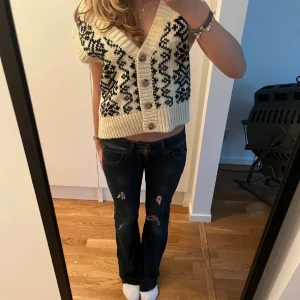 Vit stickad väst med mönster - Säljer en vit stickad väst med svart mönster och knappar framtill. Västen har en v-ringad design och är perfekt för lager-på-lager-stil. Den är kortare i modellen och passar till allt