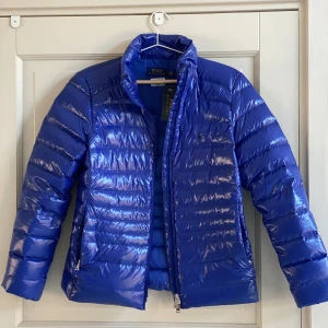 Ralph Lauren Puffer Down Jacket - Ralph Lauren Puffer Down Jacket – XS (34) – Ny med tags  Superfin och glansig dunjacka från Ralph Lauren i färgen Royal Blue. Jackan är packable (går att vika ihop i tillhörande förvaringspåse), vilket gör den perfekt för resor eller cityvardag. 	•	Helt ny, aldrig använd 	•	Med originalprislapp 	•	Storlek SP (Small Petite) – motsvarar XS / 34 	•	Glansigt material med snygg passform 	•	Kommer med matchande förvaringspåse