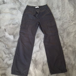 Grå/mörkblå cargobyxor med fickor - Säljer ett par grå cargobyxor med flera praktiska fickor och dragkedja. Byxorna har en loose passform och är perfekta för en avslappnad stil.