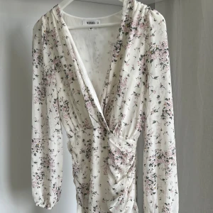 Blommig klänning från Missguided - Säljer en vacker blommig klänning från Missguided i storlek XS. Klänningen har en omlott-design med v-ringning och långa ärmar. Den är vit med ett fint blommönster i rosa och grönt. Perfekt för vår och sommar!